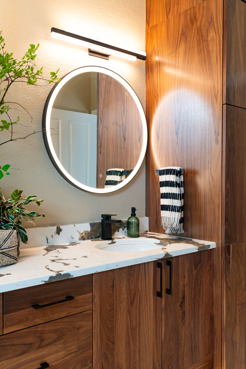 Luxury bathroom remodel featuring wood wall coverings, deep green tile work, natural accents, and a round mirror with built-in lighting for a modern, spa-inspired look.